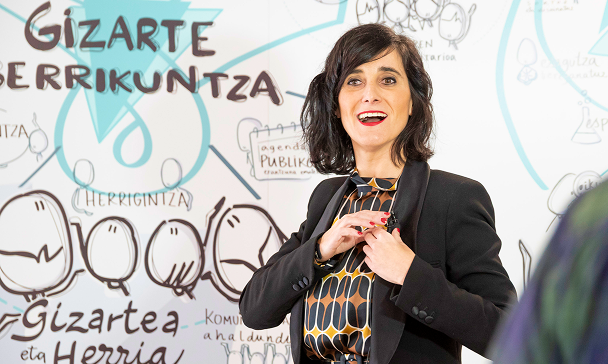 A woman in front of a board with "Gizarte berrikuntza" (social innovation) and moodboard drawings